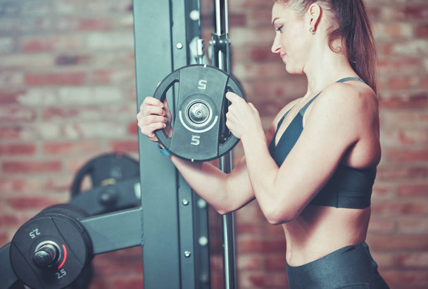 Fitness woman putting weight disk to barbell  against brick wall in gym - Photo, Image