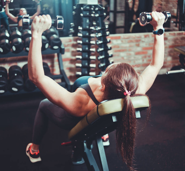 Athletic woman in sportswear doing a dumbbell press on inclined bench at gym. Back vie - Photo, Image