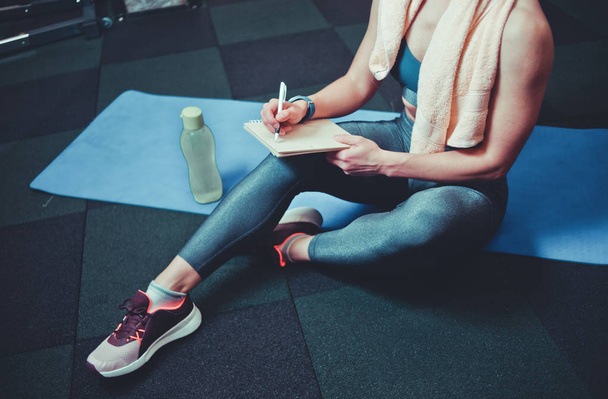 Femme sportive s'assoit sur un tapis d'entraînement et écrit de futurs plans d'entraînement pour obtenir de grands résultats en gy - Photo, image