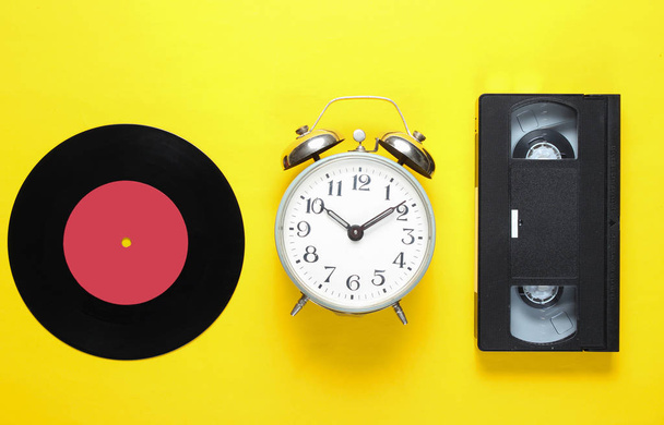 レトロなビニールレコード、古い目覚まし時計、黄色の背景にビデオテープ。80年代 トップビューミニマリズム - 写真・画像