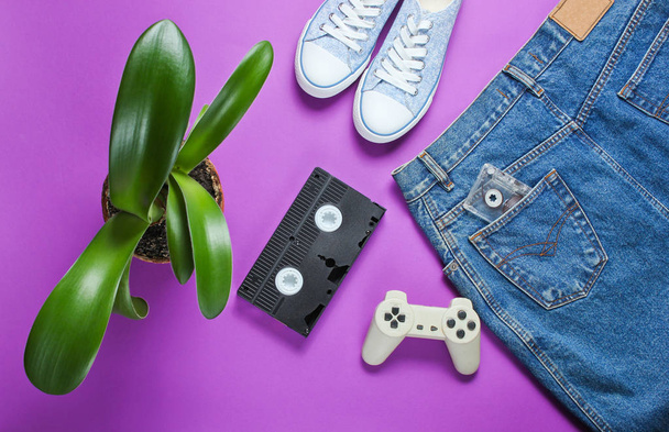 カジュアルな女性のレトロなスタイルのデニムスカート、ビデオカセット、ゲームパッド、スニーカーシューズ、紫色の背景に緑の植物とポット。ポップカルチャー、ミニマリズム - 写真・画像