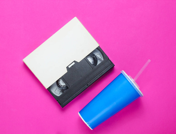 ストロー付き紙コップ、ピンクの背景にビデオカセット。ポップカルチャー80年代、レトロなスタイル。トップビュー。ミニマリズム - 写真・画像