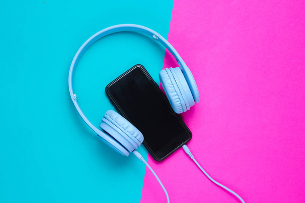 Concepto minimalista. Auriculares conectados a un teléfono inteligente en un fondo rosa-azul, vista superior
 - Foto, Imagen