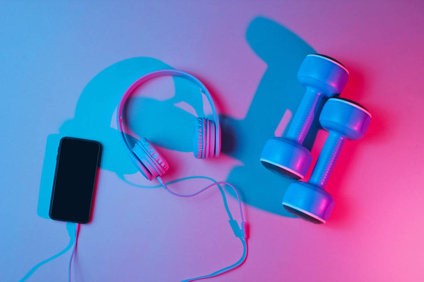 Lifestyle concept. Dumbbells with headphones and smartphone. Retro wave, blue red neon light, ultraviolet. Top view, minimalism - 写真・画像
