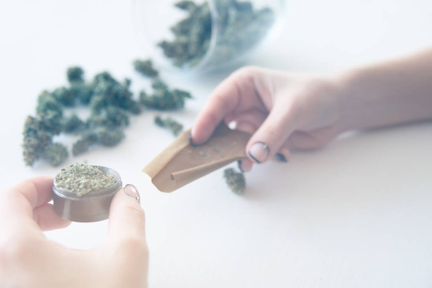 Marijuana use concept. Woman rolling a marijuana joint on white background. Woman preparing and rolling marijuana cannabis joint. Close up of marijuana blunt with grinder. Color toning - Foto, Bild
