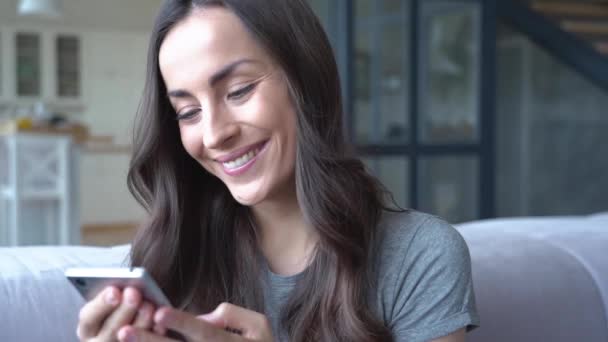 Smiling brunette woman holding smartphone while sitting on sofa at home - Кадри, відео