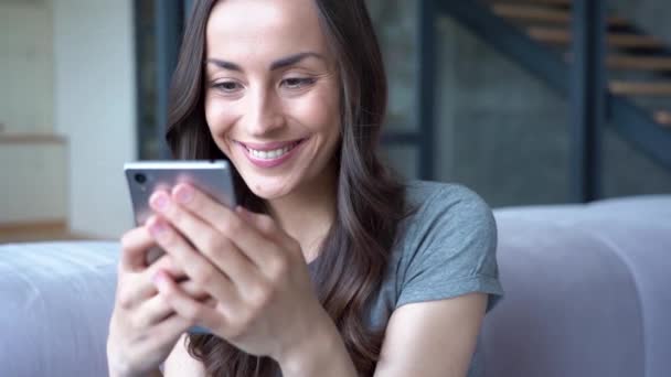 Smiling brunette woman holding smartphone while sitting on sofa at home - Filmati, video