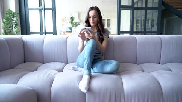 Focused young woman using smartphone while sitting on sofa - Кадри, відео