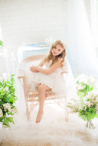 Cute little girl with blond hair in a beautiful dress in a spring studio with lilac flowers - Foto, Imagen