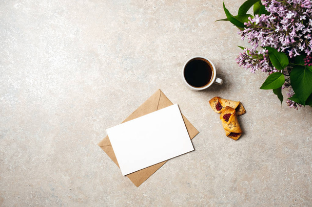 Minimal home office desk workspace with blank paper card, kraft envelope, coffee cup, cookies, lilac flowers. Flat lay, top view social media hero header background. - 写真・画像