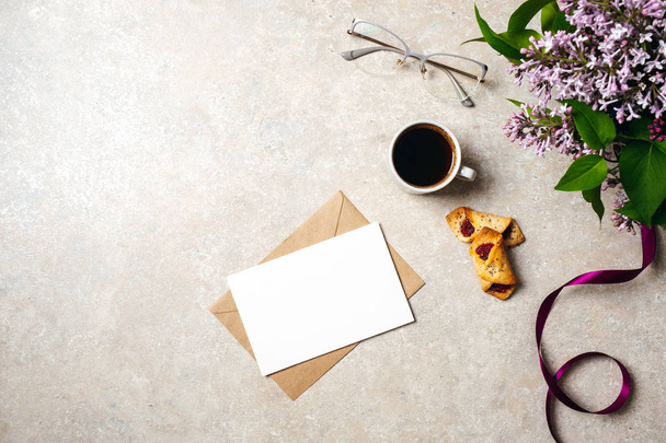 Minimal home office desk workspace with blank paper card, kraft envelope, coffee cup, cookies, lilac flowers, purple ribbon, glasses. Flat lay, top view social media hero header background. - 写真・画像