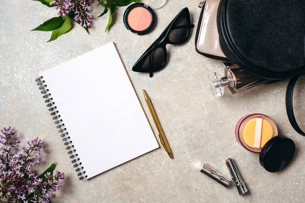 Flat lay modern minimal home workspace desk with paper notebook, golden pen, sunglasses, leather bag, wallet, woman cosmetic, spring lilac flowers on concrete stone background. Top view feminine stuff - Photo, image