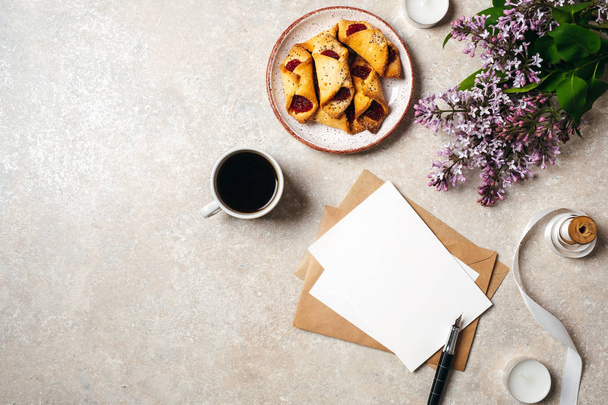 Flat lay, top view lifestyle still life composition with coffee cup, blank paper card, kraft envelope, cookies, ribbon, lilac flowers. Feminine desk with morning breakfast, cozy home concept. - 写真・画像