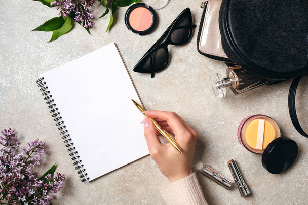 Top view hand of woman writing text message on blank paper notebook on stone table. Flat lay lilac flower, feminine cosmetic, leather bag, sunglasses. Modern minimal home workspace desk concept. - Foto, Imagen