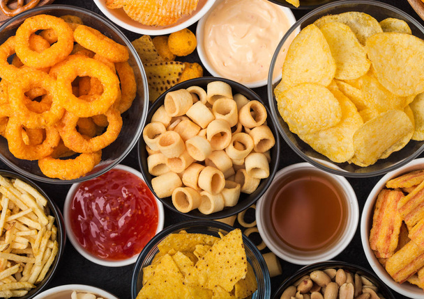 All classic potato snacks with peanuts, popcorn and onion rings and salted pretzels in bowl plates on black background. Twirls with sticks and potato chips and crisps with nachos and cheese balls. - Fotografie, Obrázek
