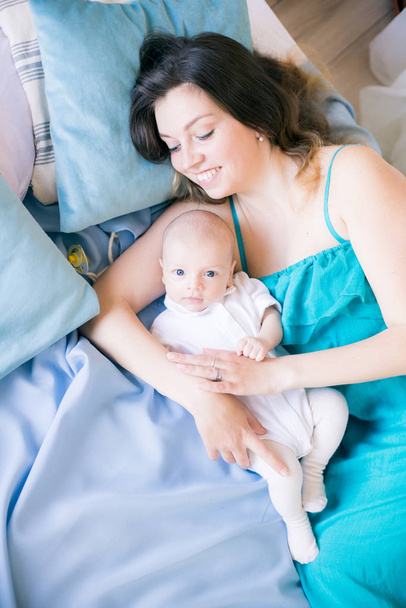 Молодая мама и маленький милый ребенок лежат на кровати с голубым одеялом дома
 - Фото, изображение