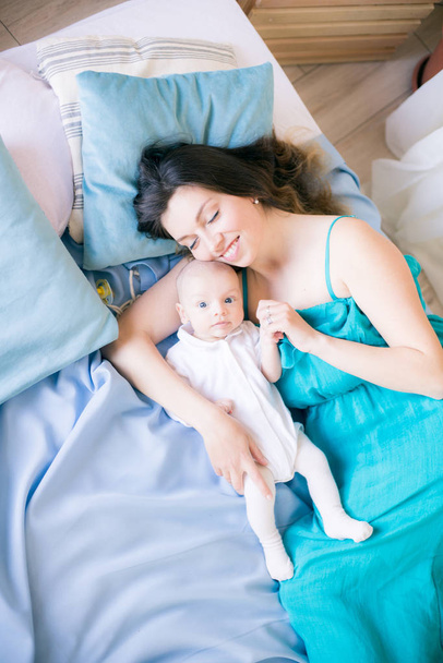 Jonge moeder en kleine schattige baby liggend op een bed met een blauwe deken thuis - Foto, afbeelding