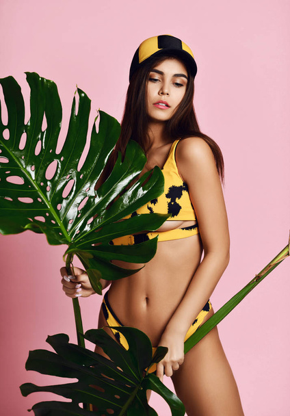 Young beautiful sexy woman with perfect smooth skin hold tropical leaves in summer hat cap and black yellow swimsuit on pink - Photo, Image