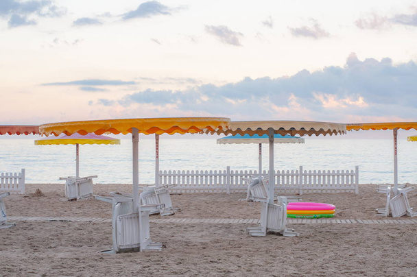 Літній морський пейзаж з білими шезлонгами під барвистими пляжними парасольками. Білий дерев'яний паркан на прозорий піщаний пляж. Яскравий рожевий дитячий басейн під пляжним парасолькою. Море відпочинок на узбережжі Чорного моря  - Фото, зображення