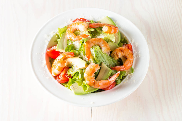 Fresh shrimp salad made of tomato, ruccola, avocado, prawns, chicken breast, arugula, crackers and spices. Caesar salad in a white, transparent bowl on wooden background - Foto, imagen