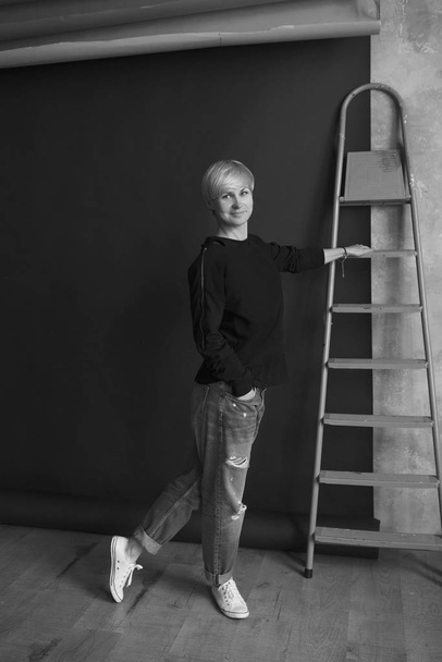 Monochrome portrait of young blonde modern woman in black sweatshirt and jeans standing near ladder (stairs) and planning renovation (repairs)  - Fotó, kép