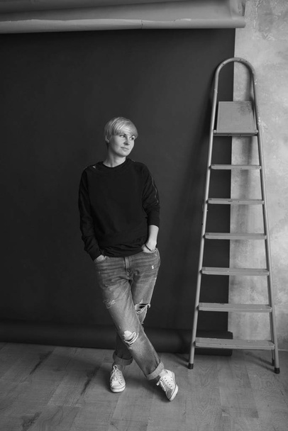 Retrato monocromático de una joven rubia moderna en sudadera negra y jeans de pie cerca de la escalera (escaleras) y planificación de renovación (reparaciones)
)  - Foto, imagen