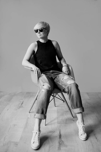 Monochrome portrait of stylish blonde young woman in black sun glasses, black shirt and jeans sitting on director chair on  wall background  - Foto, Imagen