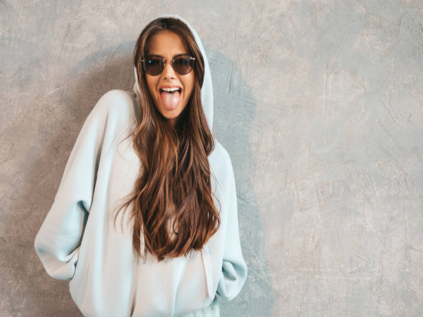 Young beautiful smiling woman looking at camera. Trendy girl in casual summer hoodie and skirt clothes. Funny and positive female posing near gray wall in studio. In sunglasses.Shows tongue - Foto, Imagen