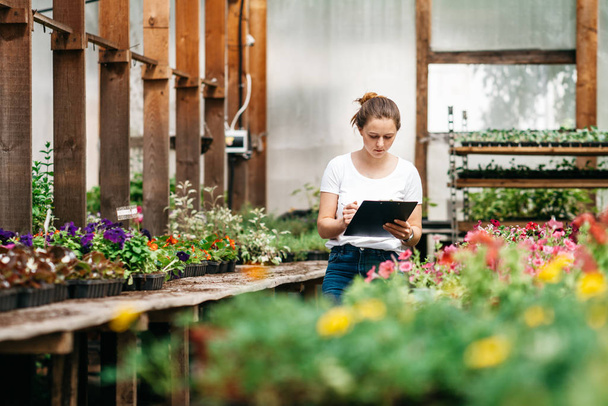 Young woman agronomist inspecting plants in greenhouse. Female gardener working in garden with flowers - Фото, зображення