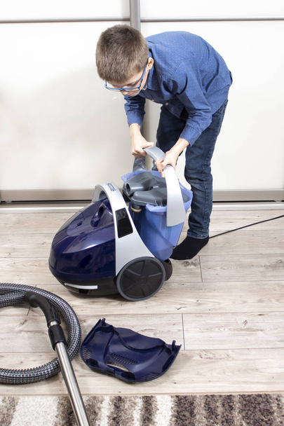 Apartment cleaning. The boy puts a container of water into the vacuum cleaner. Cleaning an apartment by a child of school age. Children's home duties. - 写真・画像