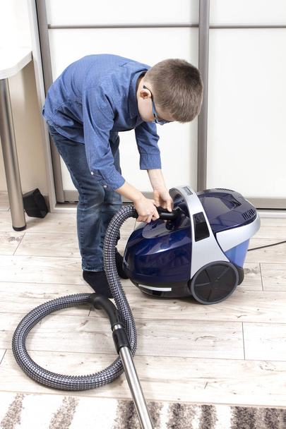 Το αγόρι ετοιμάζει μια ηλεκτρική σκούπα για να καθαρίσει το διαμέρισμα. Καθαρισμός διαμερίσματος από παιδί σχολικής ηλικίας. Παιδικά καθήκοντα στο σπίτι. - Φωτογραφία, εικόνα