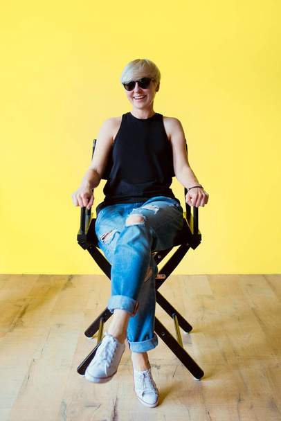 Portrait of stylish blonde young woman in black sun glasses, black shirt and jeans sitting on director chair on yellow wall background  - Photo, Image