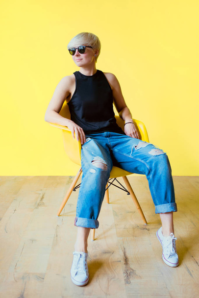 Portrait of stylish blonde young woman in black sun glasses, black shirt and jeans sitting on yellow chair on yellow wall background  - Foto, Imagen