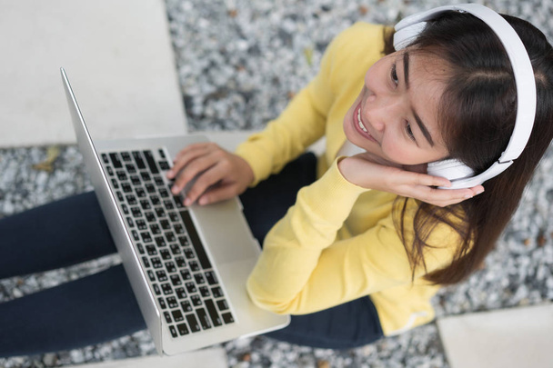 Улыбающаяся азиатская молодая девушка с ноутбуком слушает музыку по headphon
 - Фото, изображение