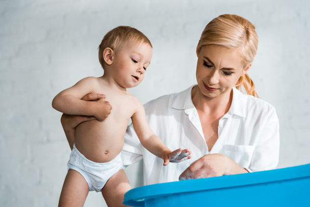 low angle view of blonde woman standing and looking at blue baby bathtub near toddler son  - Fotografie, Obrázek