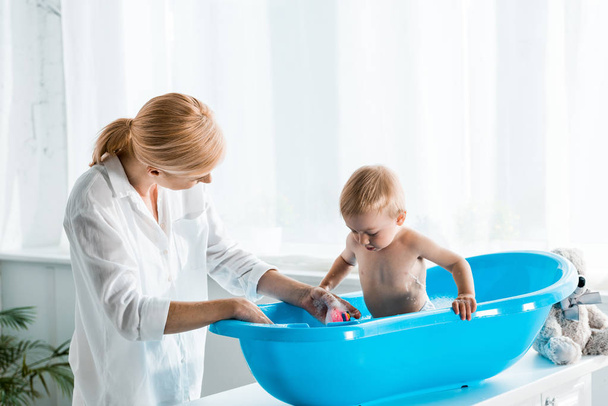 blonde mother standing near cute toddler son taking bath at home  - Photo, Image