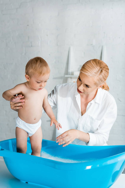 beautiful mother looking at cute toddler kid in baby bathtub  - Фото, зображення
