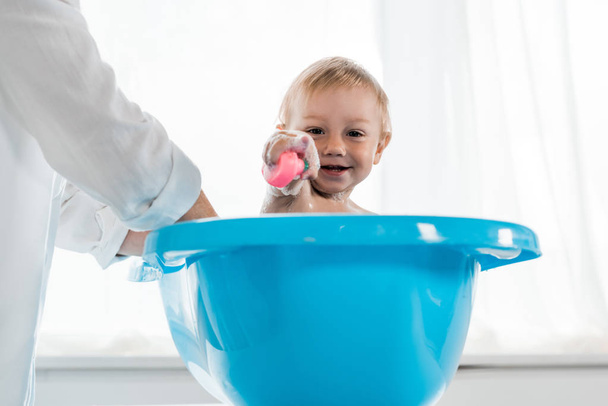Ausgeschnittene Ansicht einer Mutter, die ein glückliches Kleinkind wäscht und mit dem Finger auf die blaue Babywanne zeigt  - Foto, Bild