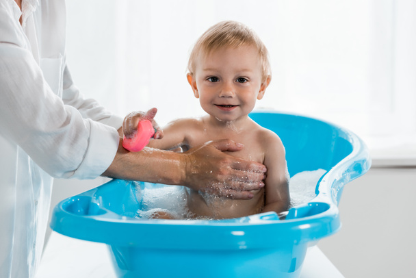 cropped view of mother washing happy toddler child with rubber toy in blue baby bathtub  - Photo, Image