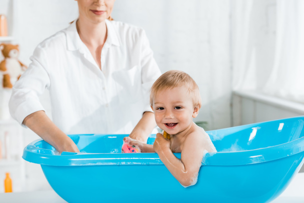 selective focus of cute smiling toddler boy in baby bathtub near mother  - 写真・画像