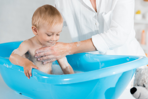 обрезанный вид на мать стирает милый малыш сын в голубой ванне ребенка
  - Фото, изображение