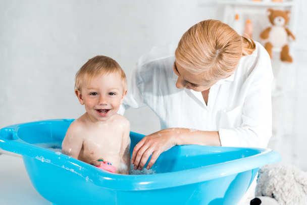blonde mother looking at cute toddler son smiling in blue baby bathtub  - Zdjęcie, obraz