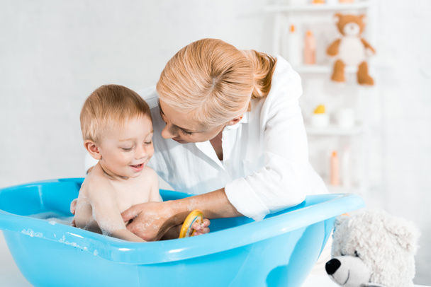 blonde mother looking at toddler son smiling in blue baby bathtub  - Zdjęcie, obraz