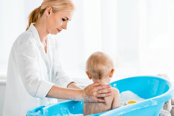 attractive mother washing and looking at toddler son in blue baby bathtub  - Photo, Image