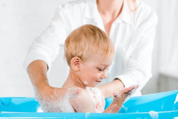 abgeschnittene Ansicht einer Frau, die niedliches Kleinkind wäscht, das Badeschaum in der Babywanne betrachtet  - Foto, Bild