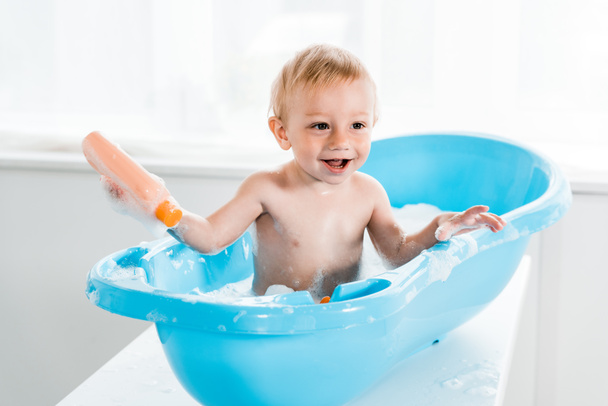 szczęśliwy Toddler dziecko uśmiechnięty podczas kąpieli w niebieskiej wanienka i trzymając butelkę z szamponem  - Zdjęcie, obraz
