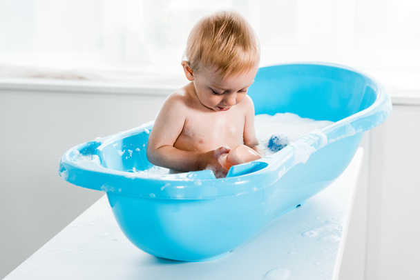 lindo niño pequeño mirando botella con champú mientras toma baño en bañera de plástico del bebé
  - Foto, imagen