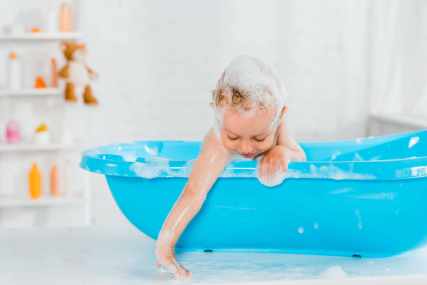 lindo niño sonriendo mientras toca la espuma del baño y tomando baño en la bañera de plástico del bebé
  - Foto, imagen