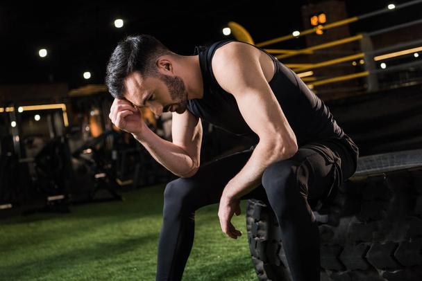 handsome and tired athletic man sitting on car tire in gym  - Photo, Image