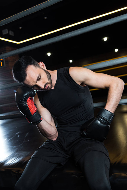 χαμηλή γωνία θέα του εξαντλημένο μούσι άνθρωπος σε μαύρο γάντια πυγμαχίας αγγίζοντας το κεφάλι στο γυμναστήριο  - Φωτογραφία, εικόνα